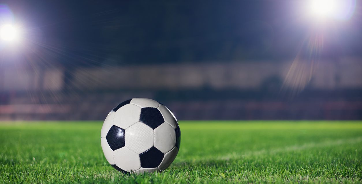 A imagem é a camera focada em uma bola de futebol, com um gramado no fundo, com duas fontes de luz da lateral superior esquerda e direita